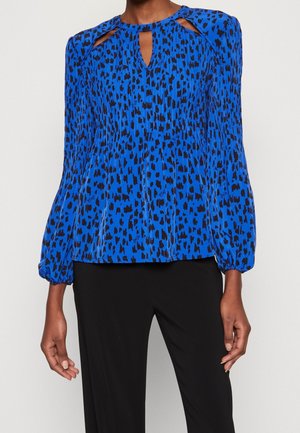 Blauwe blouse met lange mouwen en zwarte abstracte vlekken, keyhole-halslijn en elastische manchetten, gedragen met een zwarte broek door een staand persoon.