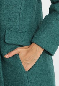 Cappotto di lana teal con finitura testurizzata, dotato di tasca laterale e design su misura. Primo piano di una mano appoggiata all'interno della tasca.
