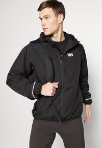 Crna lagana jakna s patentnim zatvaračem, elastičnim manžetama i porubom s uzicom. Ima bijele detalje i logo na prsima. Nosena s remenom preko ramena.