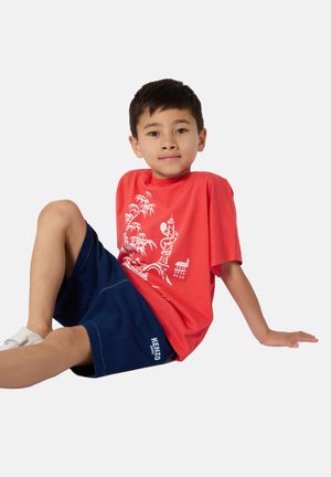 T-shirt graphique rouge avec un motif blanc, associé à un short bleu marine. Le short porte un logo brodé, et le modèle est assis.