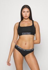 Zwarte Calvin Klein sportbeha met vierkante halslijn en logoband, gecombineerd met zwarte CK One string met logoband. Glad materiaal, getailleerd ontwerp.