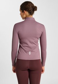 Langärmliges, eng anliegendes Oberteil aus hellmauvem Rippstoff mit hohem Kragen und weißen Chevron-Details auf der Rückseite, kombiniert mit burgunderfarbenen Leggings.