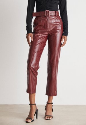 Frau trägt bordeauxrote, strukturierte High-Waist-Hose mit passendem Gürtel und schwarzen, knöchelhohen Sandaletten mit Absatz, steht auf neutralem Boden.