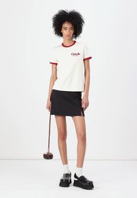 Witte t-shirt met korte mouwen, voorzien van een rode kraag en boorden, met het "Coach New York" logo. Gecombineerd met een zwarte rok en zwarte chunky schoenen.