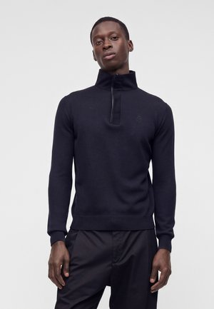 Jeune homme portant un pull noir à manches longues avec un col montant zippé au quart et un pantalon noir, debout devant un fond uni.