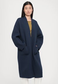 Manteau en tricot bleu marine au design long, avec des manches larges, de profondes poches et un col en V, fabriqué dans un matériau doux et texturé.
