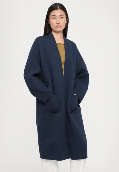 Jūras zila džemperis-mētelis ar garu dizainu, platām piedurknēm, dziļām kabatām un V veida dekoltē, izgatavots no mīksta, teksturēta materiāla.