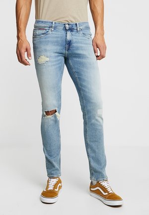 Slim Fit hellblaue Jeans mit abgenutzten Patches am Oberschenkel und Knie, getragen mit braunen und weißen Schnürsneakern.