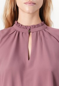 Donna che indossa una blusa mauve con colletto alto arricciato e piccolo bottone dorato al collo a fessura, vista parziale dal mento al petto.