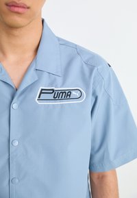Homme portant une chemise bleu clair à manches courtes avec col ouvert et écusson du logo Puma cousu sur la poitrine gauche.