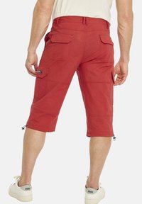 Rote Cargo-Shorts mit mehreren Taschen, einem verstellbaren Saum und einem strukturierten Stoff. Kombiniert mit weißen Sneakers, die das Markenlogo zeigen.