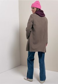 Brun uldfrakke, afslappet fit, sidelommer, båret over en mørk hættetrøje, parret med blå jeans og hvide sneakers. Lys pink strikket hue.