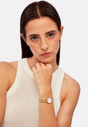 Montre bracelet en or avec un cadran rond blanc et un bracelet en métal texturé ; personne portant un top sans manches blanc, mettant en valeur la montre.