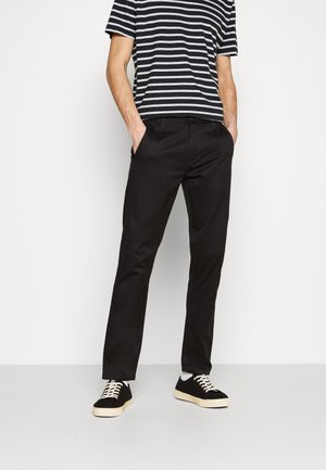 Mand iført sorte slim-fit bukser, sort-hvid stribet t-shirt, sorte sneakers med hvide snørebånd, stående med hænderne i lommerne.