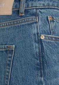 Jeans de mezclilla con una textura azul desgastada, costuras naranjas contrastantes, un parche de cuero y bolsillos visibles con detalles de remache metálico.