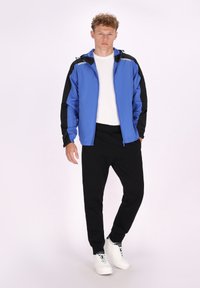 Giacca leggera blu con accenti neri, chiusura con zip davanti, polsini elasticizzati e cappuccio. Abbinata a pantaloni neri e sneakers bianche.
