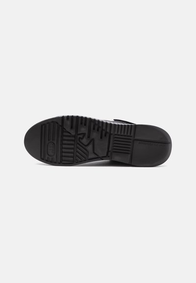 Semelle de chaussure noire avec des motifs de relief texturés variés conçus pour l'adhérence et la stabilité, vue par le dessous sur un fond uni.