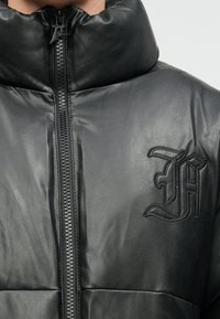 Crna kožna jakna s punjenjem, visokim ovratnikom, prednjim patentnim zatvaračem i utisnutim logotipom. Glatka tekstura s podstavljenim dijelovima za izolaciju.