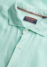 Camicia di lino verde chiaro con bottoni e colletto, con etichetta visibile del marchio Superdry all'interno della scollatura.