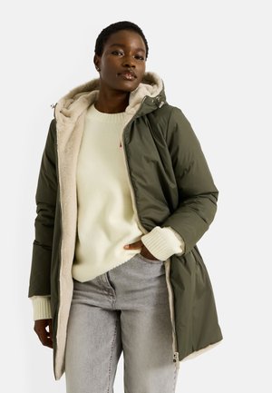 Abrigo acolchado color verde oliva con un suave forro beige, que cuenta con capucha, cierre de cremallera y bolsillos delanteros. Combinado con un suéter ligero y jeans grises.