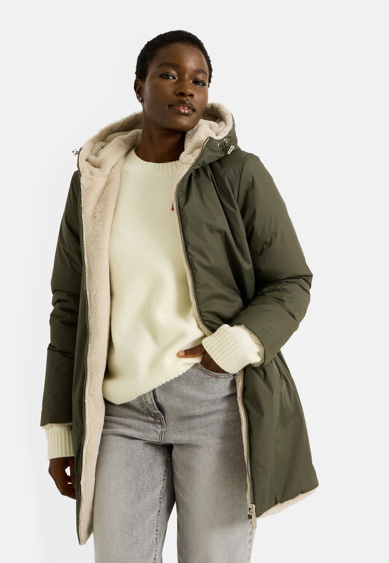 Abrigo acolchado color verde oliva con un suave forro beige, que cuenta con capucha, cierre de cremallera y bolsillos delanteros. Combinado con un suéter ligero y jeans grises.