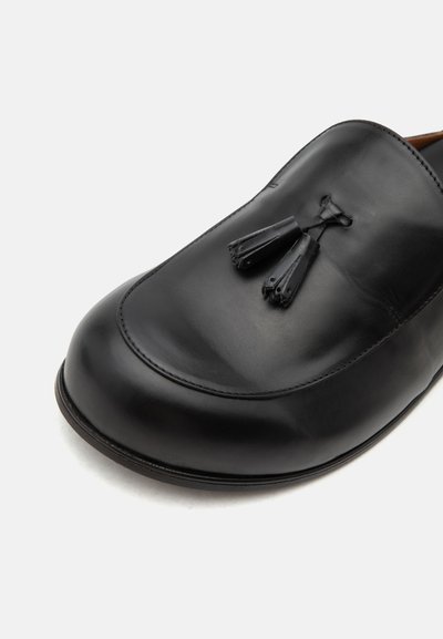 Μαύρο δερμάτινο loafers με στρογγυλεμένη μύτη και διακοσμητικά φουντάκια στην κορυφή, φωτογραφημένο σε λευκό φόντο.