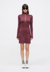 Mauve långärmad klänning med hög krage och dragkedja med flerfärgad rand. Modellen bär grå strumpor och vita sneakers.