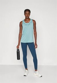 Hele sinine sportlik tanktop logoga, kombineeritud tumesiniste retuusidega; modell hoiab musta joogipudelit ja kannab valgeid tenniseid.