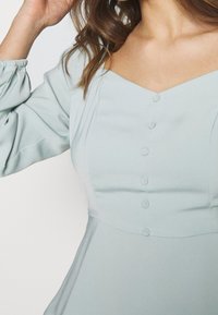 Abito verde chiaro con scollo a V, caratterizzato da una parte anteriore con bottoni, maniche arricciate e una texture liscia. Pieghe decorative sul corpetto.