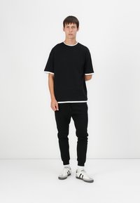 Schwarzes kurzärmeliges Hemd mit weißem Rand, kombiniert mit schwarzen Jogginghosen. Das Outfit hat einen lässigen Schnitt und wird durch weiße Sneaker ergänzt.