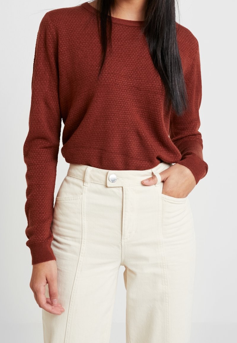 Femme portant un haut à manches longues texturé couleur rouille, rentrée dans un pantalon crème taille haute, avec une main dans la poche.