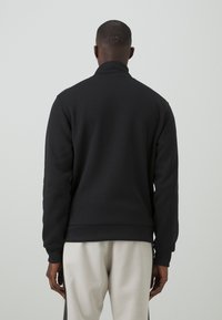 Czarny sweatshirt z wysokim kołnierzem, ściągaczami na mankietach i teksturowanym wykończeniem. Styl jest casualowy i dopasowany w talii, zestawiony z jasnymi spodniami.