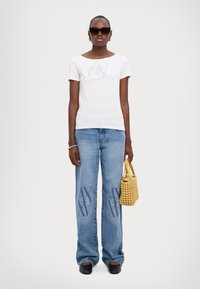 Camiseta blanca de manga corta con estampados de logo, combinada con jeans de pierna ancha de color azul claro que presentan un patrón. Accesorios con un bolso tejido amarillo.