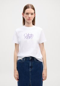 T-shirt en coton blanc avec le logo "Calvin Klein" en cursive violette, porté avec une jupe en denim bleu foncé, texture lisse, design à col rond.