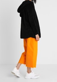 Sort, hætteklædt, fuzzy jakke, kombineret med lys orange cropped bukser. Hvide sneakers med grå detaljer. Enkel, afslappet pasform generelt.