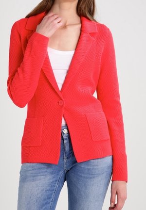 Femme portant un blazer rouge vif texturé sur un haut blanc et un jean bleu clair, avec une main touchant son cou.
