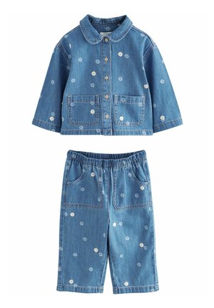 Enfant portant une veste et un pantalon en denim bleu avec de petits motifs de fleurs blanches et des boutons marron, debout avec la main gauche détendue.