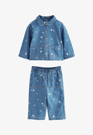 Dziecko w niebieskiej dżinsowej kurtce i spodniach z małymi białymi kwiatowymi nadrukami i brązowymi guzikami, stoi z lewą ręką w spoczynku.