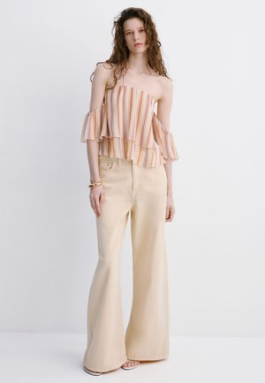 Femeie purtând un top în dungi cu volane și umeri goi și pantaloni bej largi, stând în fața unui fundal alb simplu.