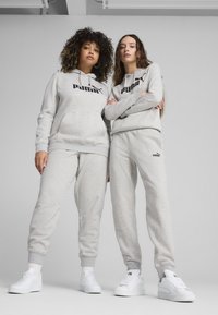 Zwei Modelle tragen passende hellgraue Kapuzenpullover und Jogginghosen mit dem schwarzen Puma-Logo. Weiße Sneakers vervollständigen den lässigen Look.