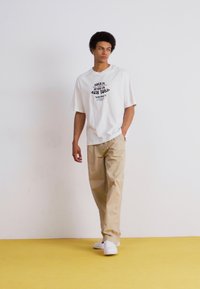 Hvid t-shirt med sort tekstdesign, parret med beige bukser og hvide sneakers. Enkelt, afslappet pasform. Stående mod en neutral baggrund.