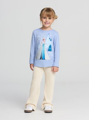 Ragazza giovane sorridente con capelli biondi indossa una maglia a maniche lunghe di colore azzurro chiaro a tema Frozen, pantaloni a coste color crema e sneaker bianche su sfondo grigio.