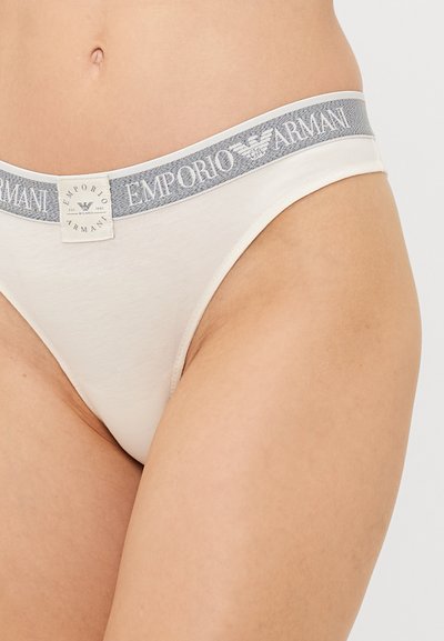 Beige cotton thong with a gray elastic waistband featuring "Emporio Armani" logo and eagle emblem. Smooth texture, minimal coverage design.