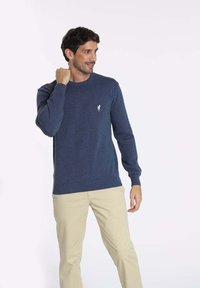 Pull bleu marine en tricot avec un col rond, des poignets et un ourlet côtelés, arborant un petit logo blanc sur la poitrine.