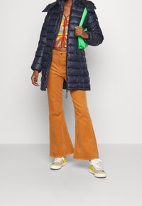 Marinblå puffjacka, orange kordfloddsbyxor med utsvängda ben, mönstrad crop top, grön handväska och sneakers med gula detaljer.