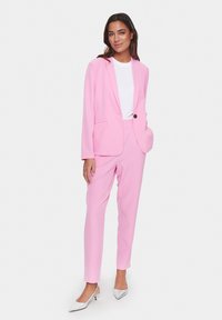 Rosa Anzug, bestehend aus einem Blazer mit Taschen und passenden Hosen, aus glattem Stoff gefertigt. Getragen mit einem weißen Hemd und silbernen High Heels.