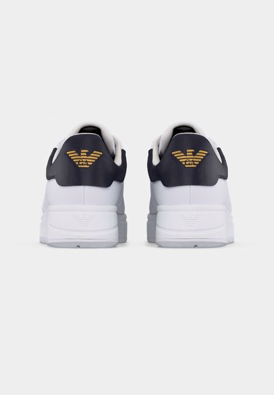 Λευκά αθλητικά παπούτσια με μαύρες ταμπέλες στη φτέρνα που φέρουν χρυσά λογότυπα αετών, απεικονιζόμενα από πίσω σε ένα απλό ανοιχτό φόντο.