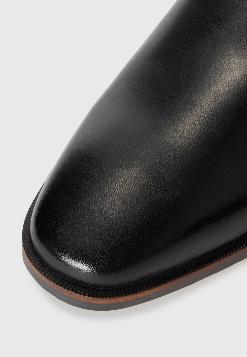 Zbliżenie na elegancki czarny skórzany czubek buta z gładkim wykończeniem i brązową warstwową podeszwą na białym tle.