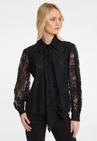 Schwarze Spitzenbluse mit langen, durchsichtigen Ärmeln, einem spitzen Kragen und einem gebundenen Detail vorne, versehen mit floralen Mustern und einer fließenden Silhouette.