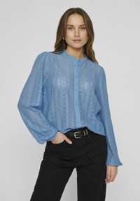 Blouse en dentelle bleu clair avec un col rond, des boutons à l'avant et des manches longues. Présente des motifs délicats et une coupe décontractée. Associée à un pantalon noir.